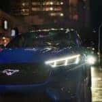 A person is shown walking from a blue 2024 Ford Mustang Mach-E GT SUV parked on a city curb.
