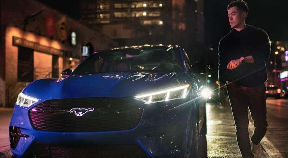 A person is shown walking from a blue 2024 Ford Mustang Mach-E GT SUV parked on a city curb.
