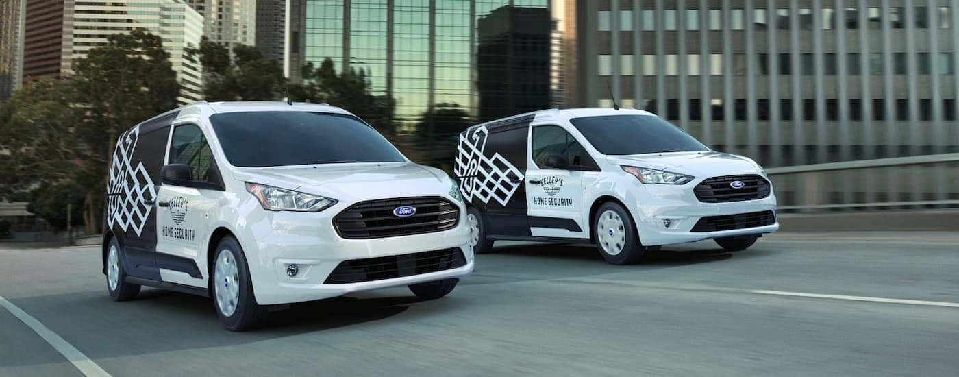 Two white and black 2023 Ford Transit Connects are driving through a city.