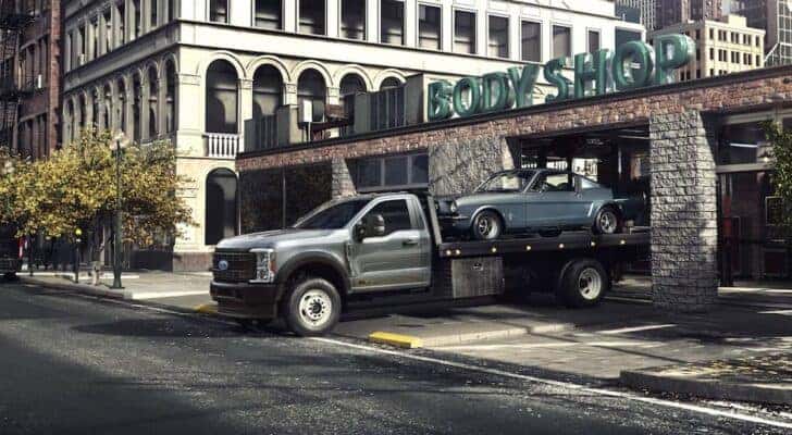 A grey 2024 Ford F-600 Super Duty Chassis Can is leaving a body shop with a blue vehicle.