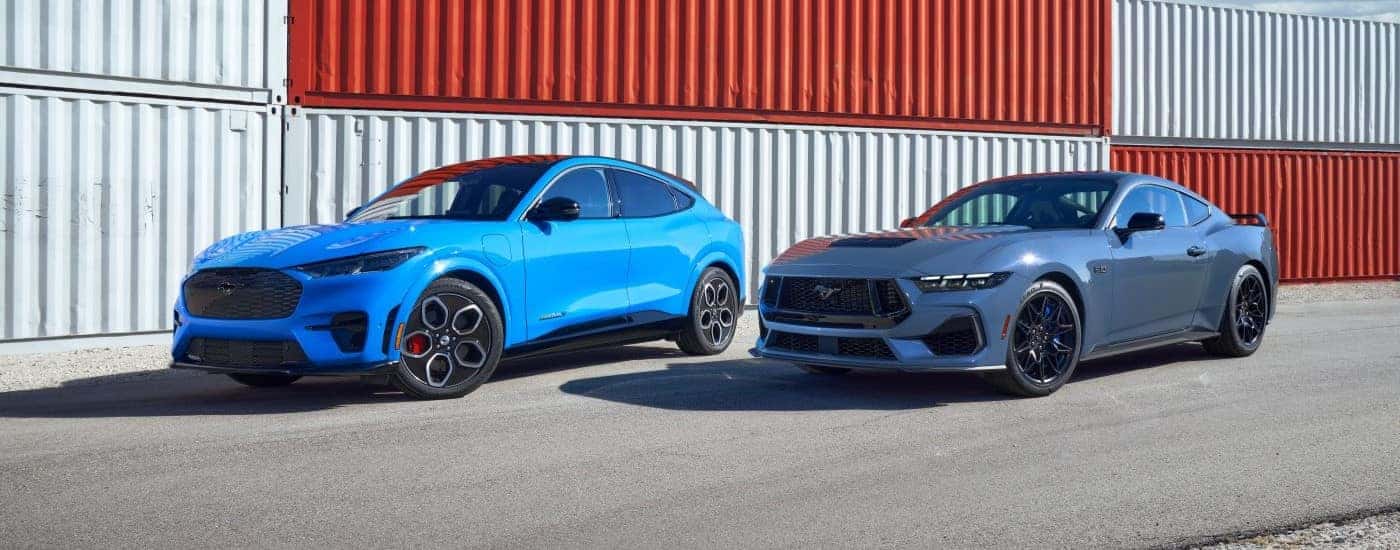 A blue 2024 Ford Mustang Mach-E and a blue Mustang are shown passing shipping containers.