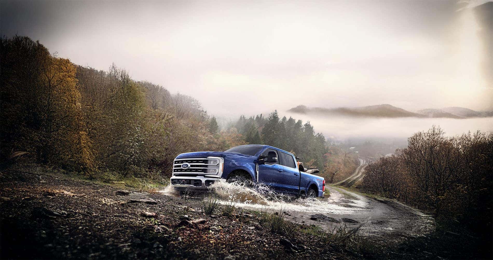 A 2026 Ford Super Duty driving off‑road in West Virginia, kicking up dust as it climbs a rugged trail surrounded by trees and rolling hills under a bright sky.