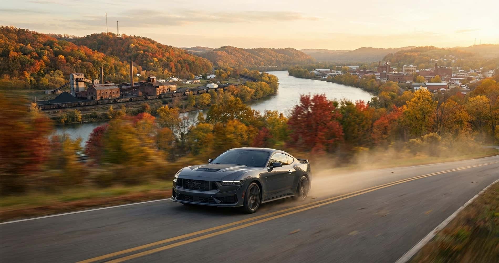2026 Ford Mustang driving on a two lane road in the mountains with a river and small rural town in the distance surrounded by vibrant autumn foliage