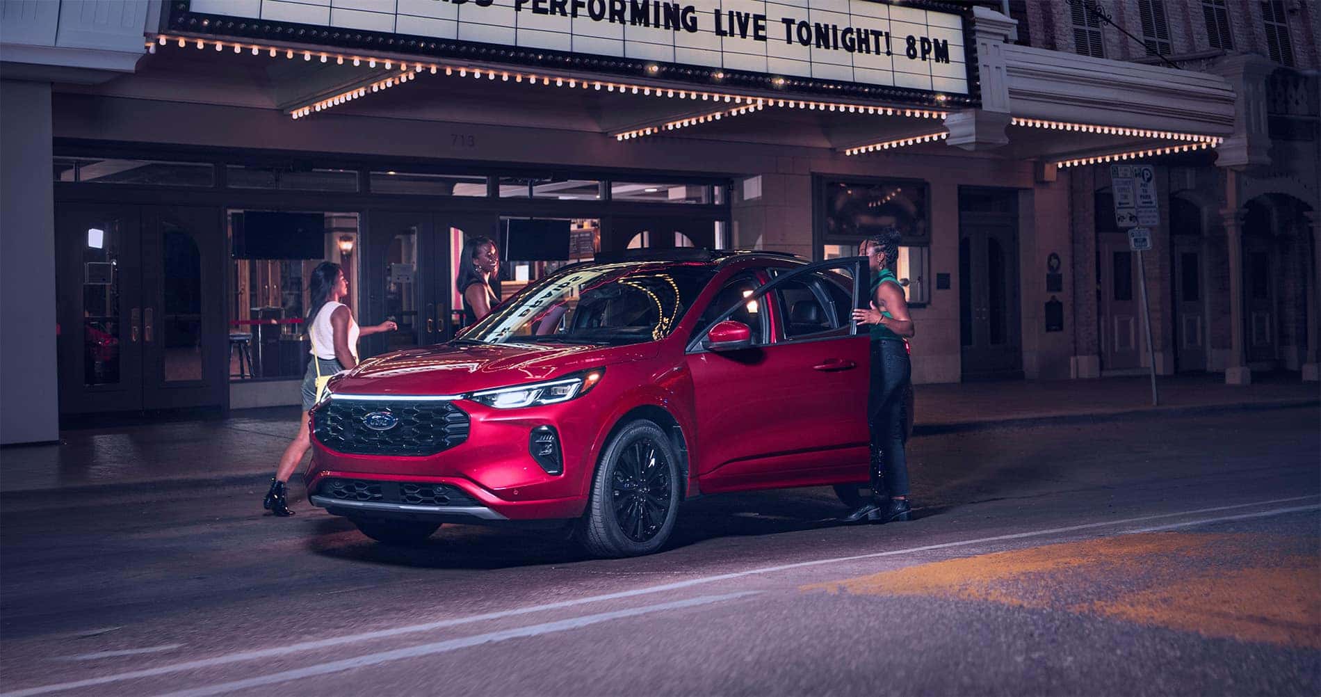 A red Ford Escape ST Line SUV parked at night in front of a theater with a glowing marquee reading “Performing Live Tonight 8PM,” with three people gathered around the vehicle as warm lights from the entrance illuminate the sidewalk and surrounding buildings.
