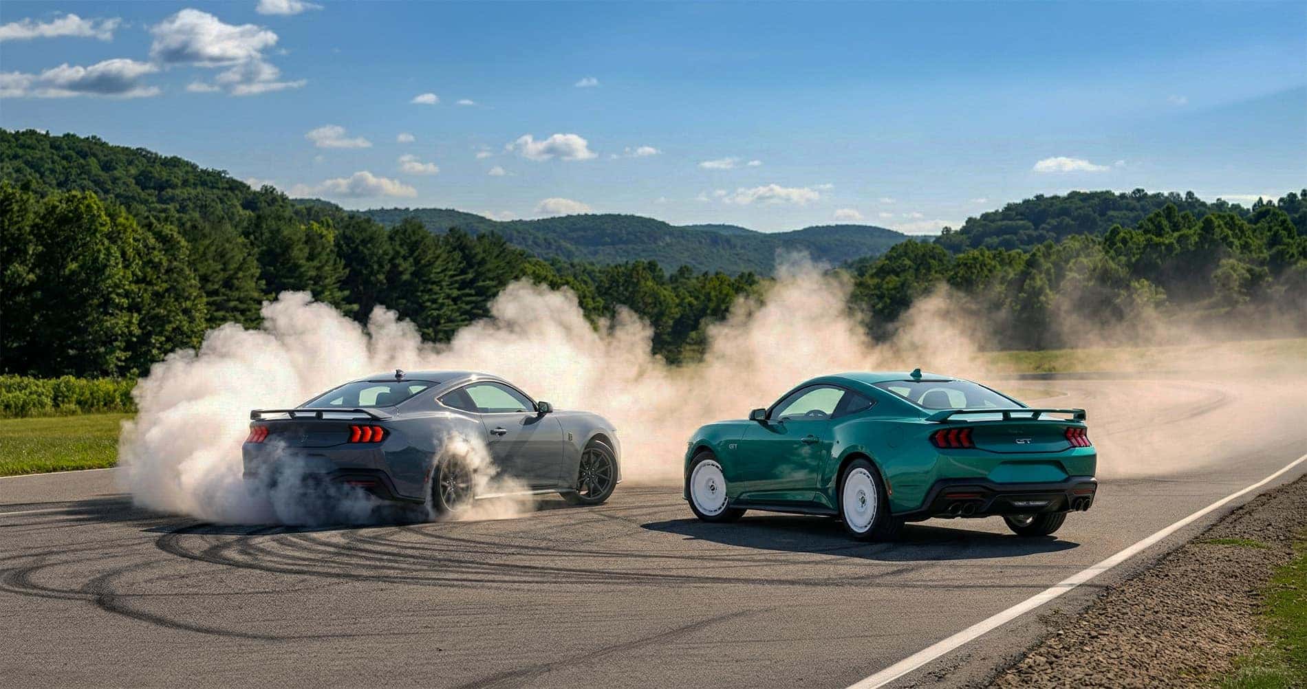 Two 2026 Ford Mustang sports cars on a racetrack, one gray doing a burnout and one green that is parked, with thick tire smoke billowing behind them, surrounded by green trees and hills under a bright sky.