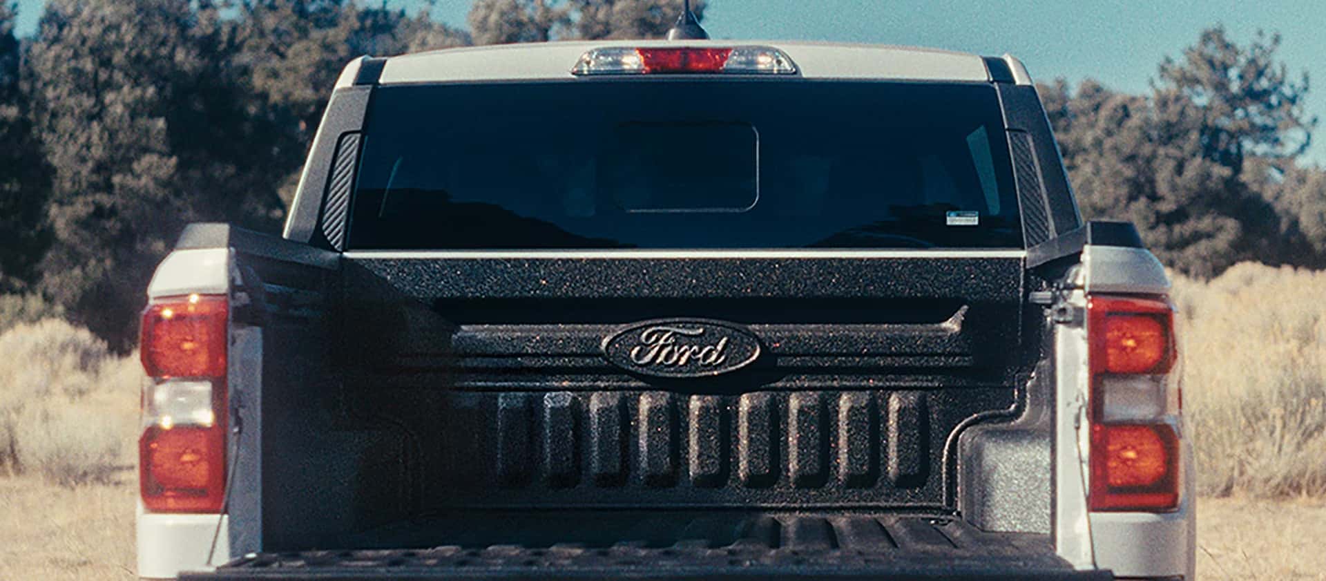 Rear view of a 2026 Ford Maverick with the tailgate down, showing the empty bed with a durable liner, parked outdoors in a dry, open landscape with trees in the background