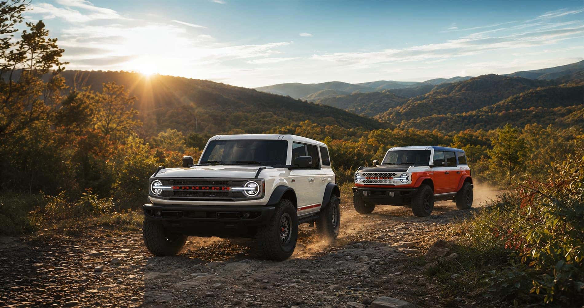 Two 2026 Ford Bronco special-edition SUVs drive along a rocky mountain trail near White Hall, West Virginia, with trees, rolling hills, and morning sunlight in the background.