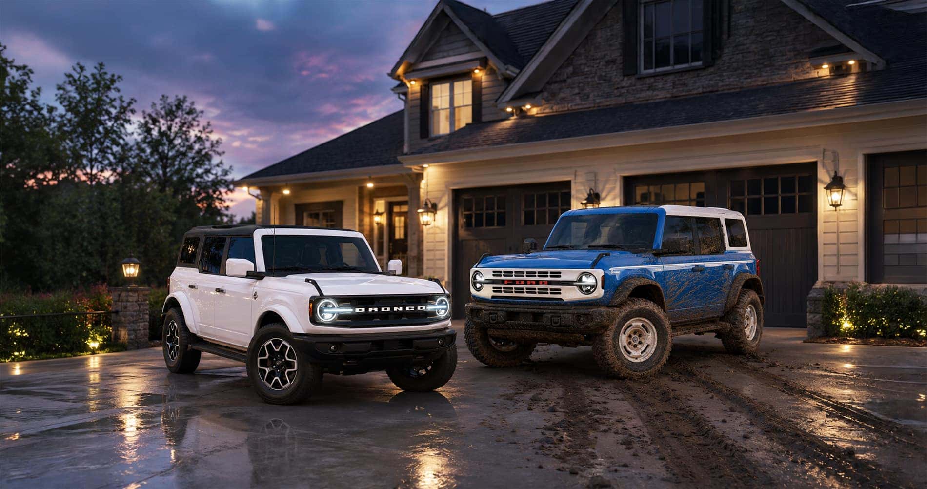 Two 2026 Ford Bronco SUVs—a clean white Heritage Edition and a mud-covered blue Badlands—parked side by side in a residential driveway at dusk, with warm house lights reflecting on the wet pavement, highlighting the Bronco’s versatility for both adventure and everyday life.