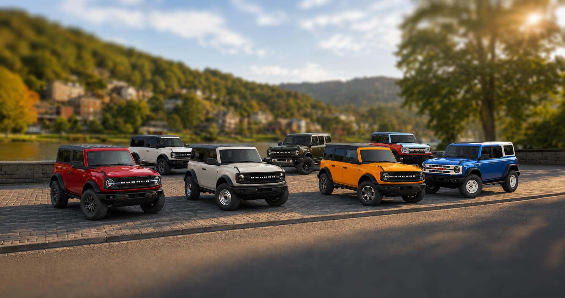 Seven 2026 Ford Bronco SUVs in red, gray, white, green, orange, and blue parked along a riverside street in Whitehall, West Virginia, with hillside homes and trees in the background under a bright morning sky, illustrating the full range of Bronco trim levels and color options.