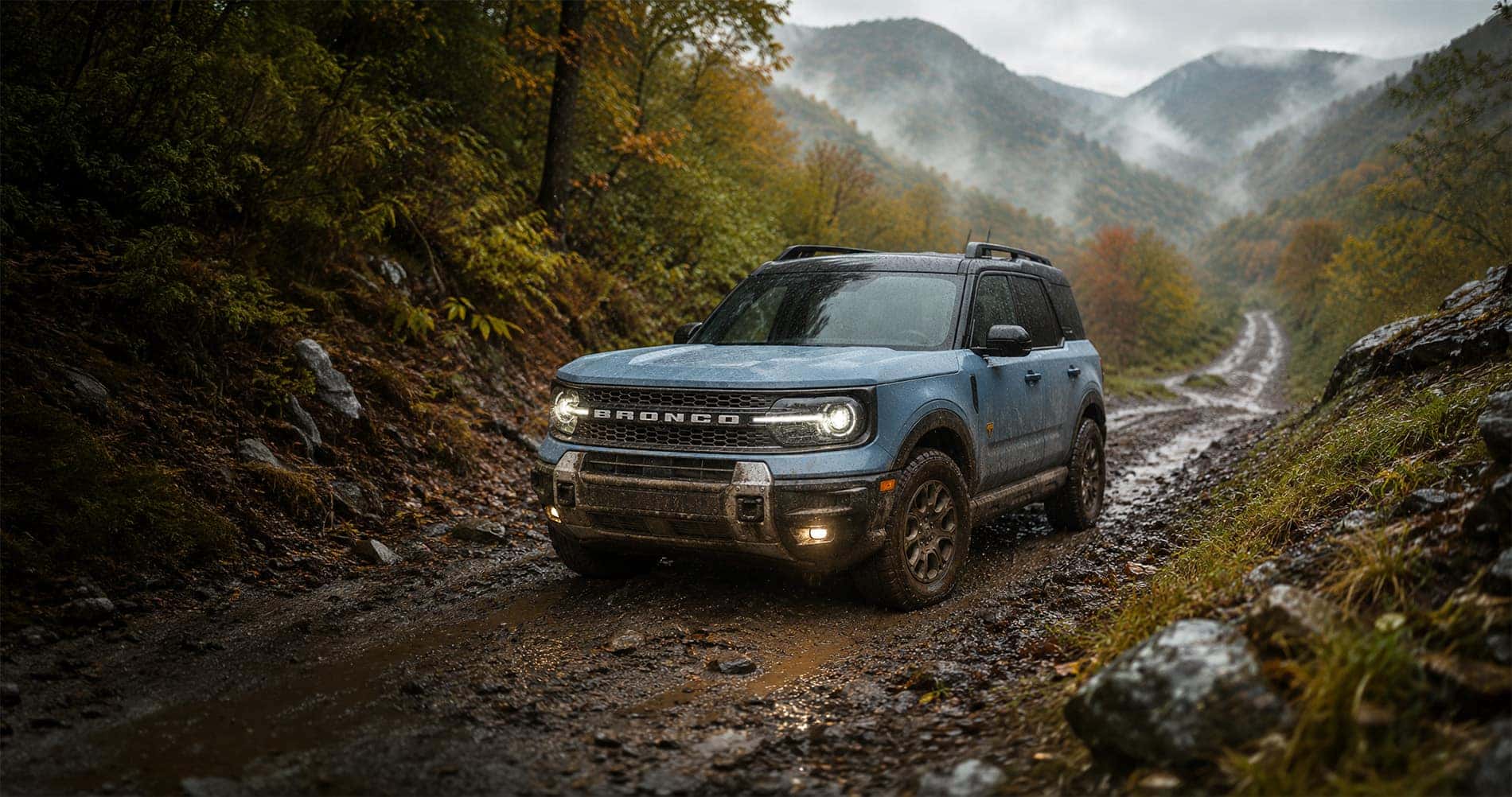 Blue Ford Bronco Sport Badlands driving on muddy mountain trail surrounded by misty forest highlighting off road capability.