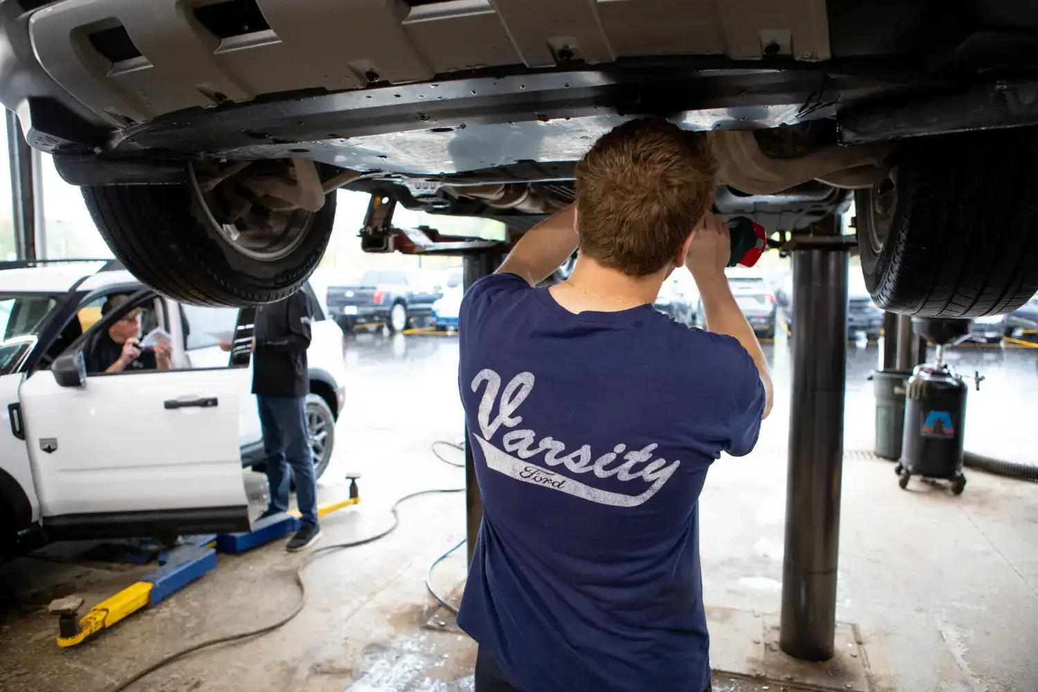 A technician in a dark blue Varsity Ford branded shirt performs maintenance on the underside of a vehicle elevated on a hydraulic lift inside a professional automotive service garage, with tools, equipment, and other cars visible in the background.