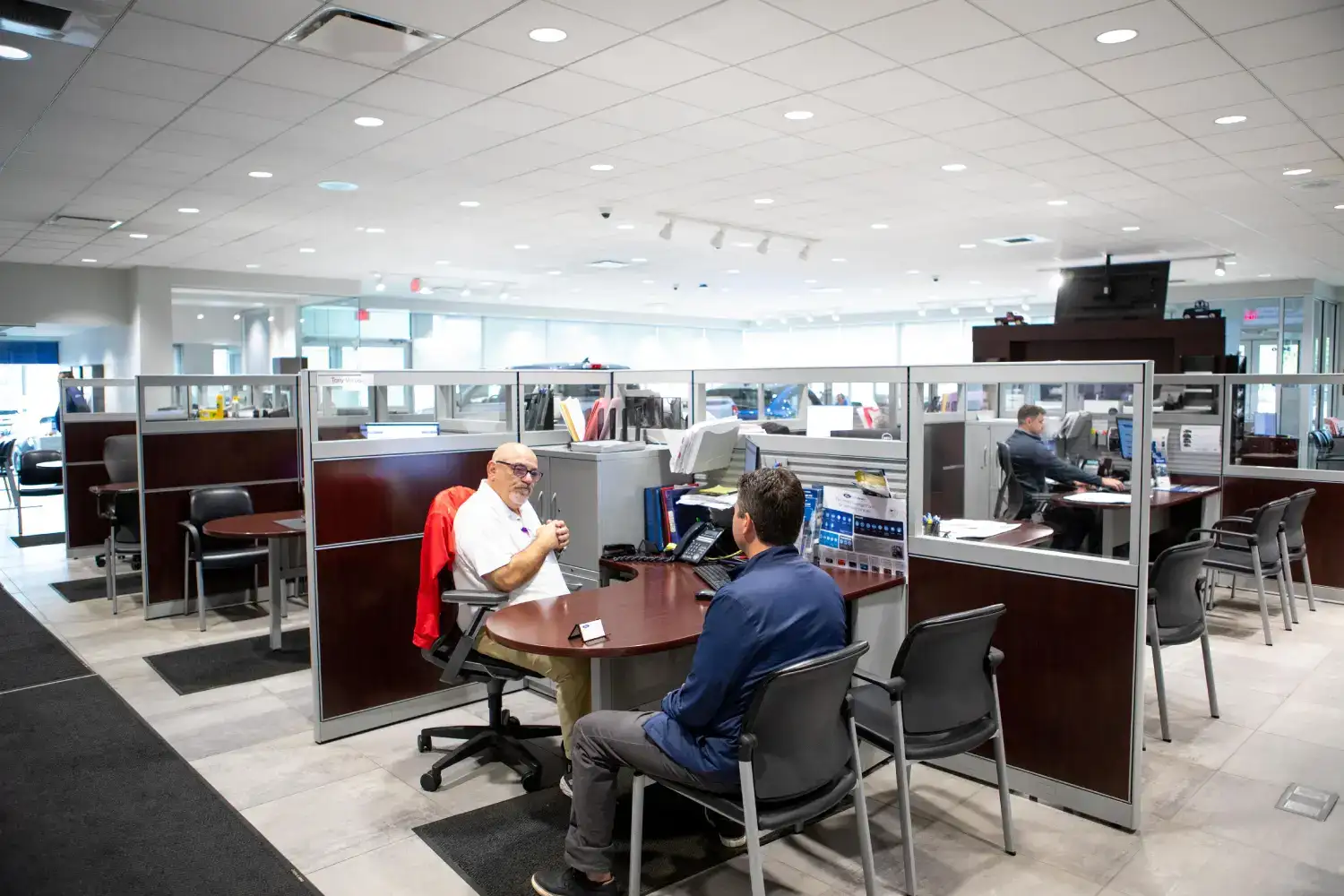 In the open sales floor of Varsity Ford, a sales consultant wearing a white shirt engages in conversation with a customer across a small round table in a semi-private cubicle, reviewing documents amid a brightly lit office environment with additional workstations in the background.