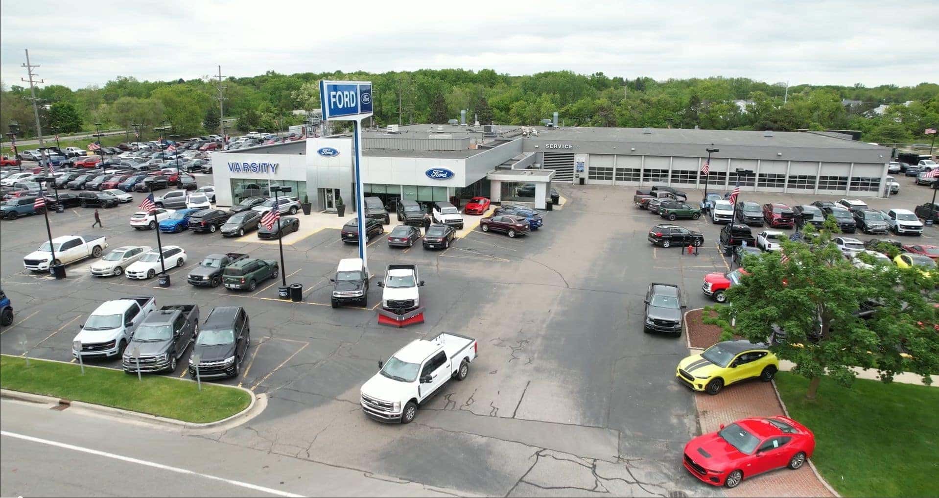 An aerial view of Varsity Ford dealership on an overcast day, showing a large parking lot filled with numerous new Ford vehicles including trucks, SUVs, and cars. The modern dealership building features prominent "VARSITY" and "Ford" signage on the facade, a tall Ford pole sign, American flags throughout the lot, and a separate service bay area to the right. Green trees surround the property in the background.