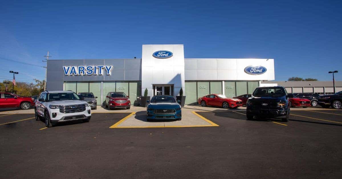 "Ford Dealer in Ann Arbor, MI" A modern Ford dealership building with a prominent "VARSITY" sign in large blue letters on the left and the Ford logo centered above the entrance. The facade features large glass windows and doors under a clear blue sky. In the foreground, three Ford vehicles are prominently displayed in the parking lot: a white SUV on the left, a teal Mustang in the center, and a black pickup truck on the right. Additional red vehicles are visible in the background lot, with American flags on poles to the sides.