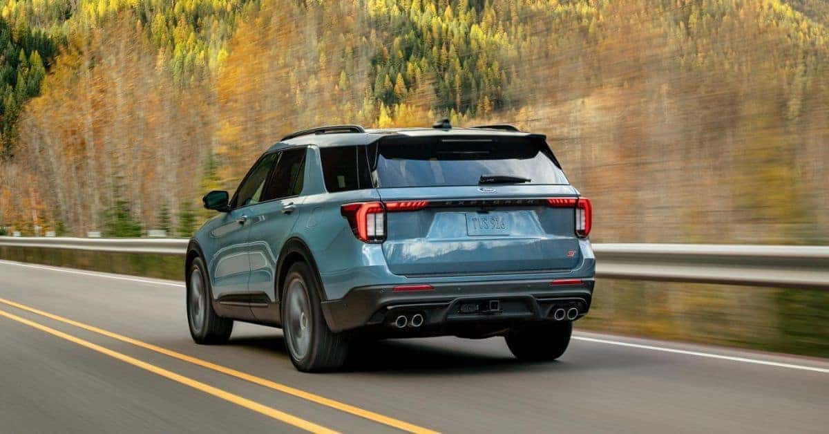 A blue Ford Explorer SUV driving on a highway from a rear three-quarter view, with autumn foliage in the background.