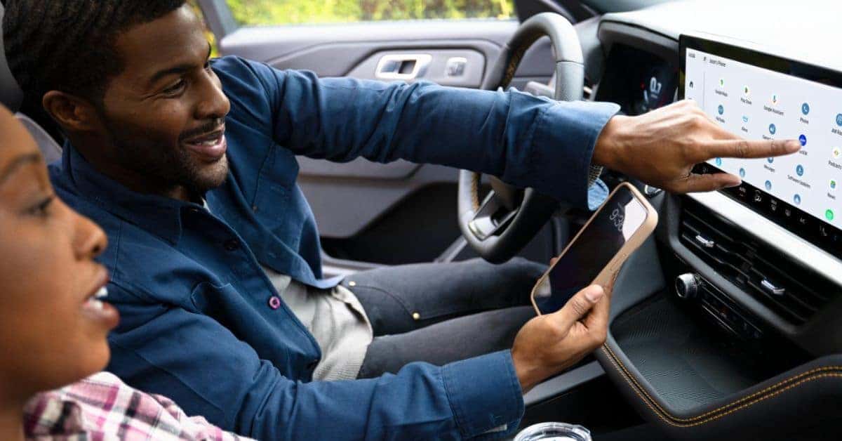A man smiling while driving a Ford Explorer, holding a phone in one hand, viewed from inside the vehicle focusing on the driver and steering wheel.