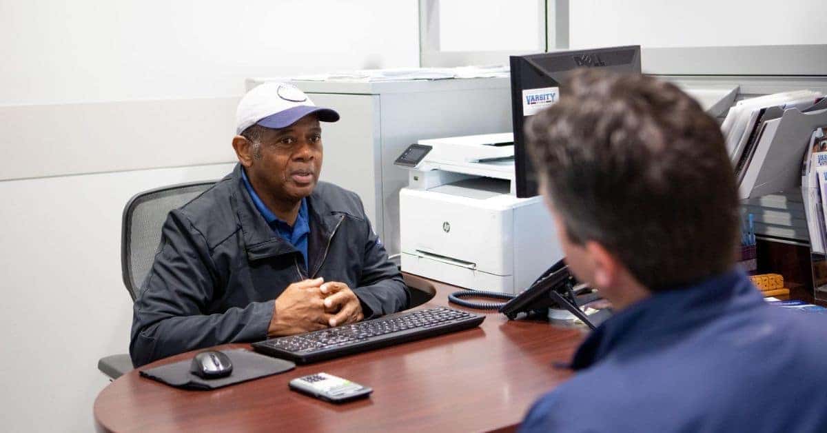 "Sales Team at Varsity Ford" Two individuals engaged in conversation at a desk in an office setting at Varsity Ford. A sales representative wearing a white and blue cap, blue shirt, and dark jacket sits with hands clasped, smiling while speaking to a customer whose back is to the camera. The desk has a computer, keyboard, mouse, phone, and various office supplies. A "VARSITY" branded sign is visible in the background.