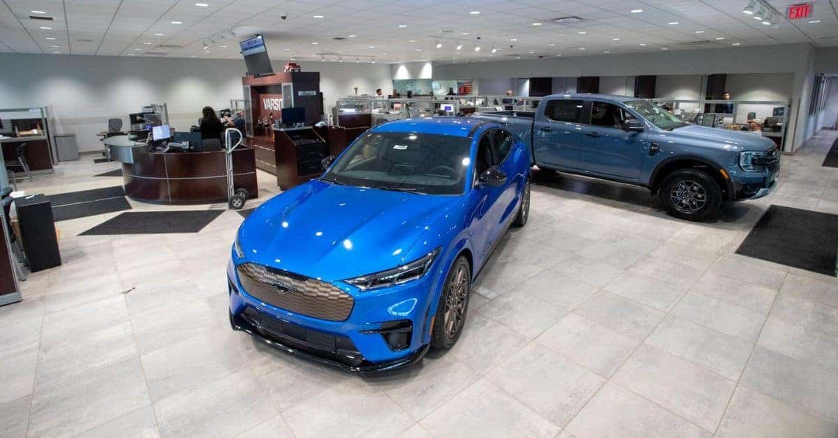 "Showroom at Varsity Ford" Interior showroom of Varsity Ford dealership featuring two blue Ford vehicles on display: a sleek electric Mustang Mach-E in the foreground and a Ford Maverick pickup truck in the background. The modern showroom has polished tile flooring, bright overhead lighting, and a reception desk with staff in the distance. A "VARSITY" sign is visible behind the counter area.