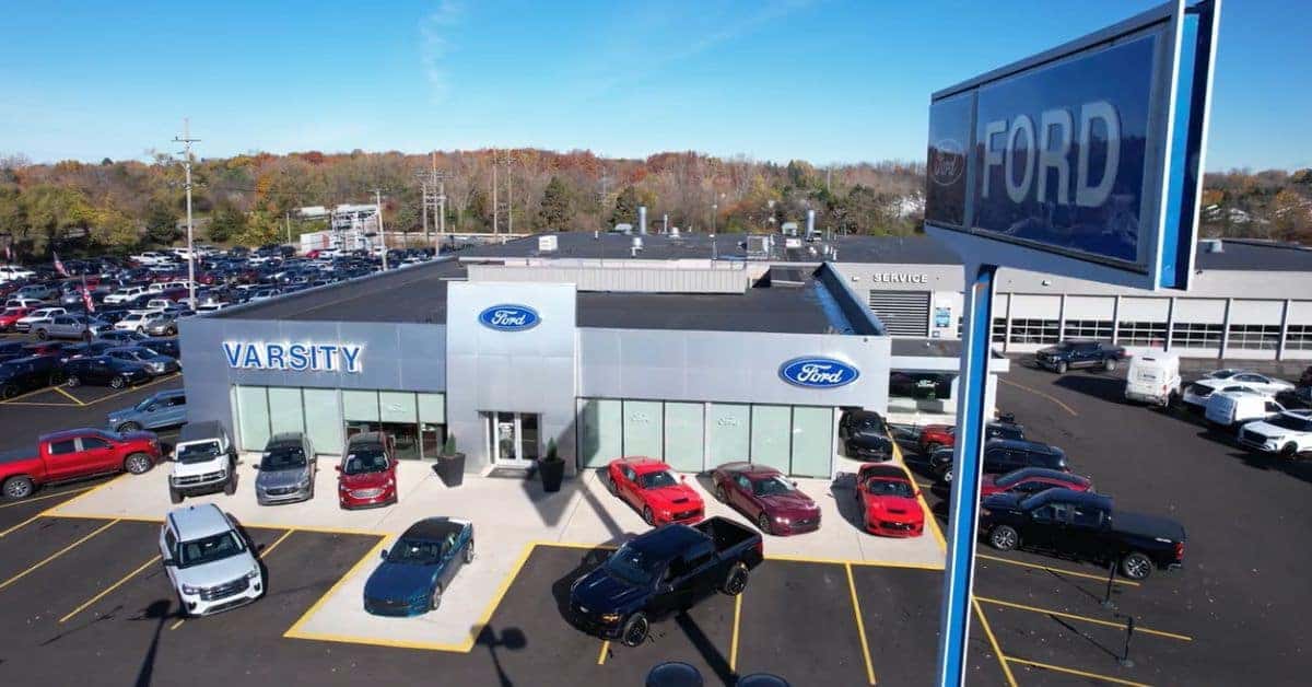 "Varsity Ford in Ann Arbor, MI" An aerial view of Varsity Ford, a modern Ford dealership, featuring a large building with prominent "VARSITY" signage in blue on the left and Ford logos on the facade. The main entrance has large glass doors and windows, with several Ford vehicles displayed in front, including red Mustangs, a black pickup truck, a blue sedan, and others. The parking lot is filled with numerous cars, extending to the left and rear. A tall Ford sign is visible in the foreground on the right, and autumn-colored trees line the background under a partly cloudy sky. A "SERVICE" entrance is noted on the right side of the building.