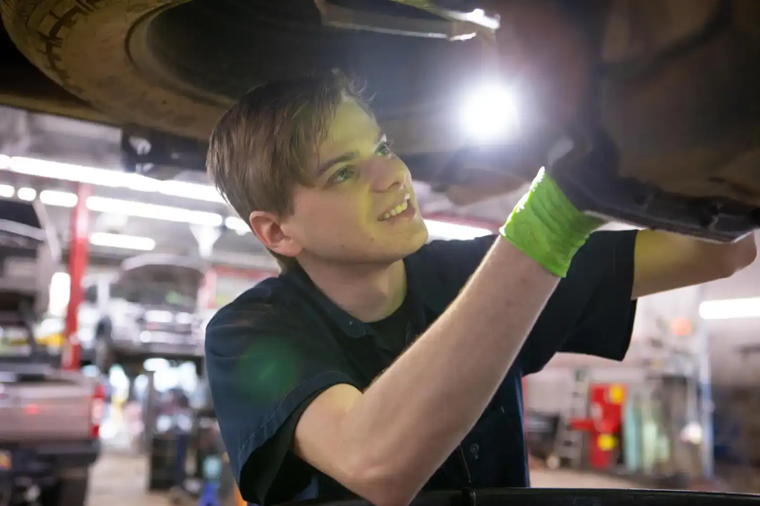 A young automotive technician in a dark short-sleeved uniform and bright green gloves works underneath a raised vehicle, holding a flashlight to illuminate the undercarriage while smiling toward the camera in a brightly lit service garage with blurred vehicles and equipment in the background.