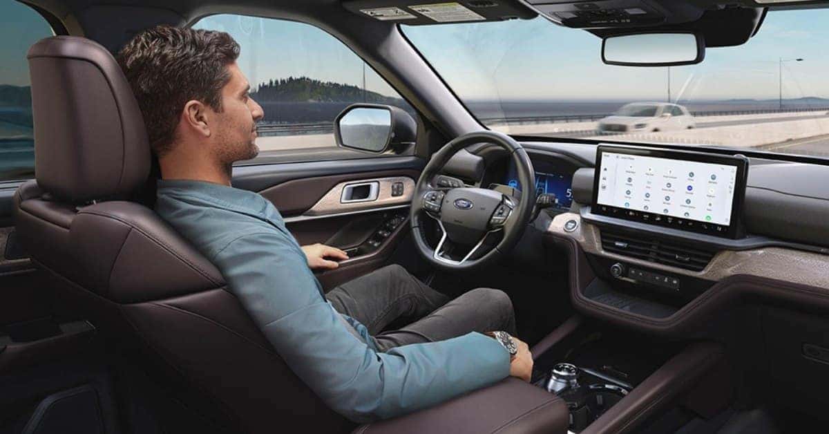 Interior view of a modern SUV from the back seat, showcasing a driver, a large infotainment screen, and a sophisticated dashboard design. The vehicle is in motion on a highway with a scenic landscape in the background. A man is driving hands-free using the Ford BlueCruise.