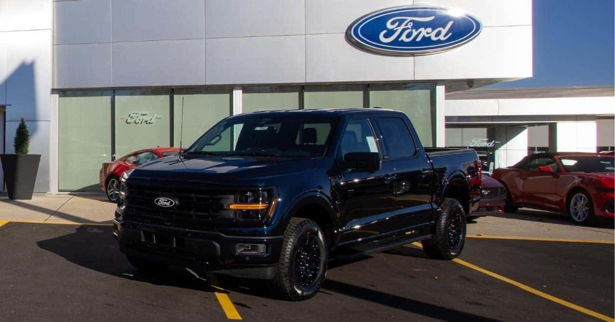 A dark blue 2026 Ford F-150 pickup truck parked in front of a modern Ford dealership lot, with a red sports car visible and a large Ford logo on the building facade.