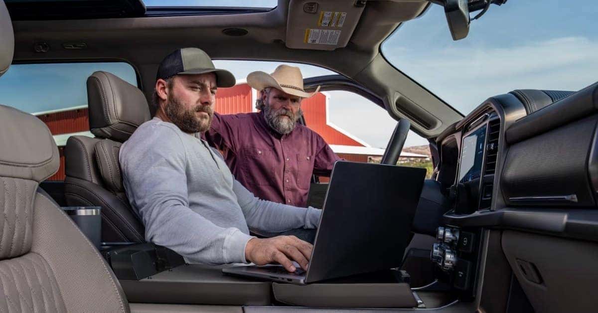 Interior of a modern 2026 Ford F-150 pickup truck with two men, one using a laptop on a fold-out console. The truck features luxurious leather seats, a large infotainment screen, and a rural farm setting visible through the windows.