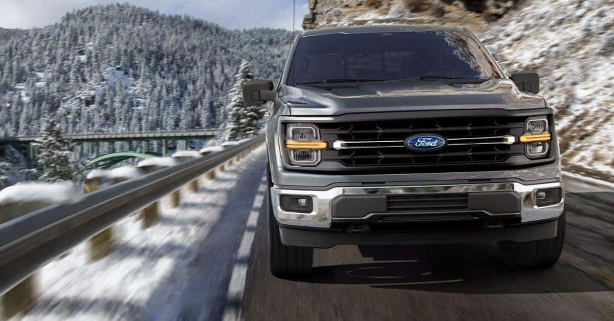 A gray 2026 Ford F-150 pickup truck driving on a snow-covered mountain road, with motion blur emphasizing its speed. The background features snow-covered pine trees, a green bridge spanning a valley, and towering snowy mountains under an overcast sky.
