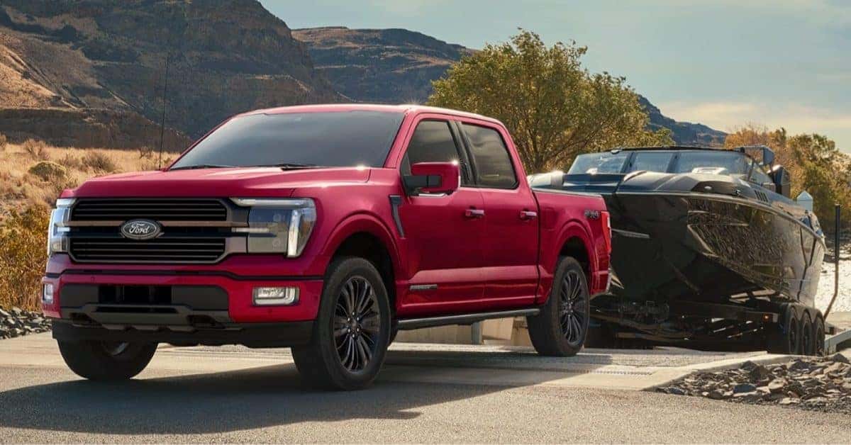 A red 2026 Ford F-150 pickup truck towing a black speedboat on a trailer, parked near a boat ramp with a mountainous landscape and clear blue sky in the background.