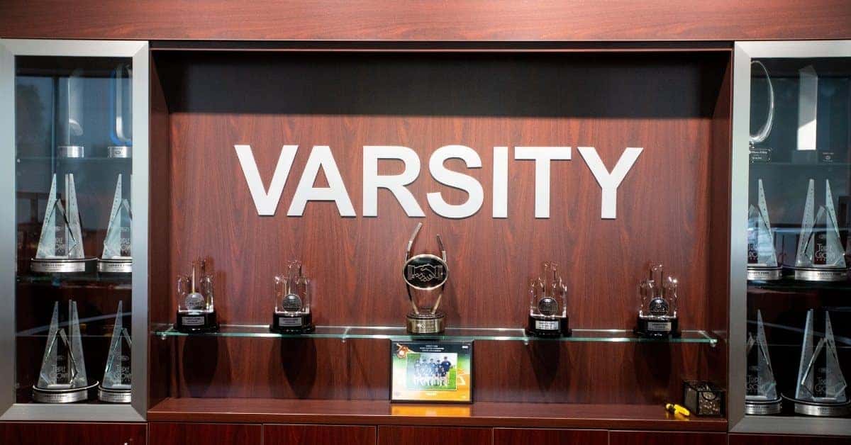 A wooden trophy display case with a central 'VARSITY' sign, featuring various awards including a 'Dealer of the Year 2021' trophy, surrounded by symmetrical glass cabinets holding pyramid-shaped trophies.