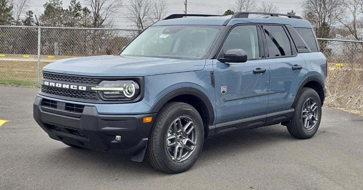 A light blue 2026 Ford Bronco Sport SUV parked on a paved surface, featuring a bold front grille with 'BRONCO' lettering, round LED headlights, and rugged black trim, set against an overcast sky and a chain-link fence.