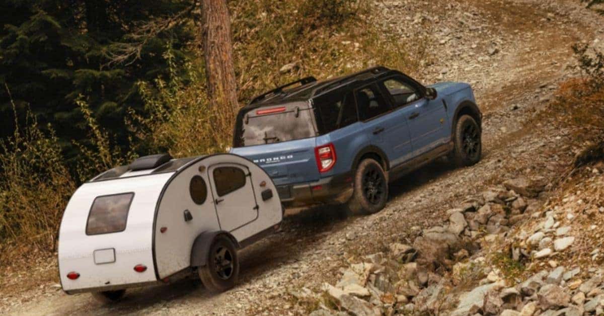 A blue 2026 Ford Bronco Sport SUV towing a white teardrop camper trailer uphill on a rocky dirt road surrounded by pine trees and dry brush, showcasing its off-road capability and adventurous design.