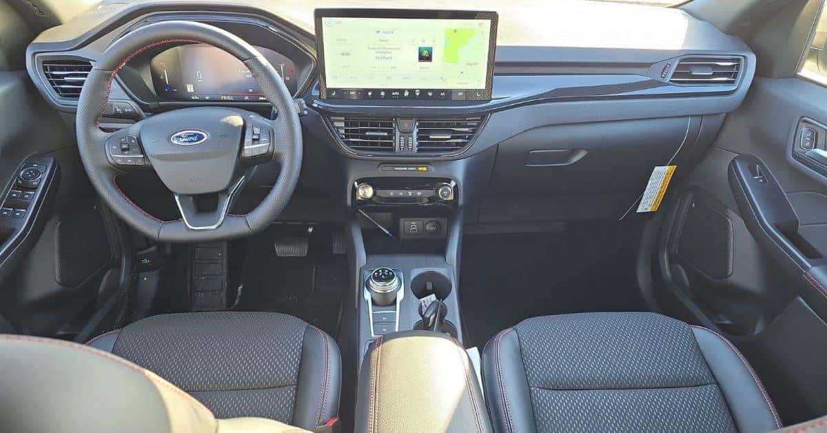 Interior of a 2026 Ford Escape highlighting the dashboard, steering wheel with red stitching, digital instrument cluster, and a large infotainment screen displaying SiriusXM and navigation. The center console includes climate controls, USB ports, and a rotary gear selector.