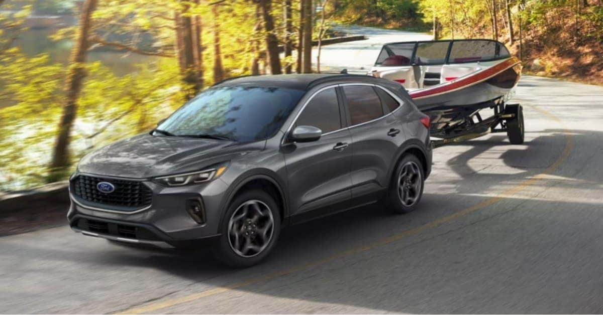 A dark grey 2026 Ford Escape towing a white and red boat on a trailer along a winding road surrounded by trees and a body of water. The scene conveys outdoor adventure and the vehicle's towing capability.