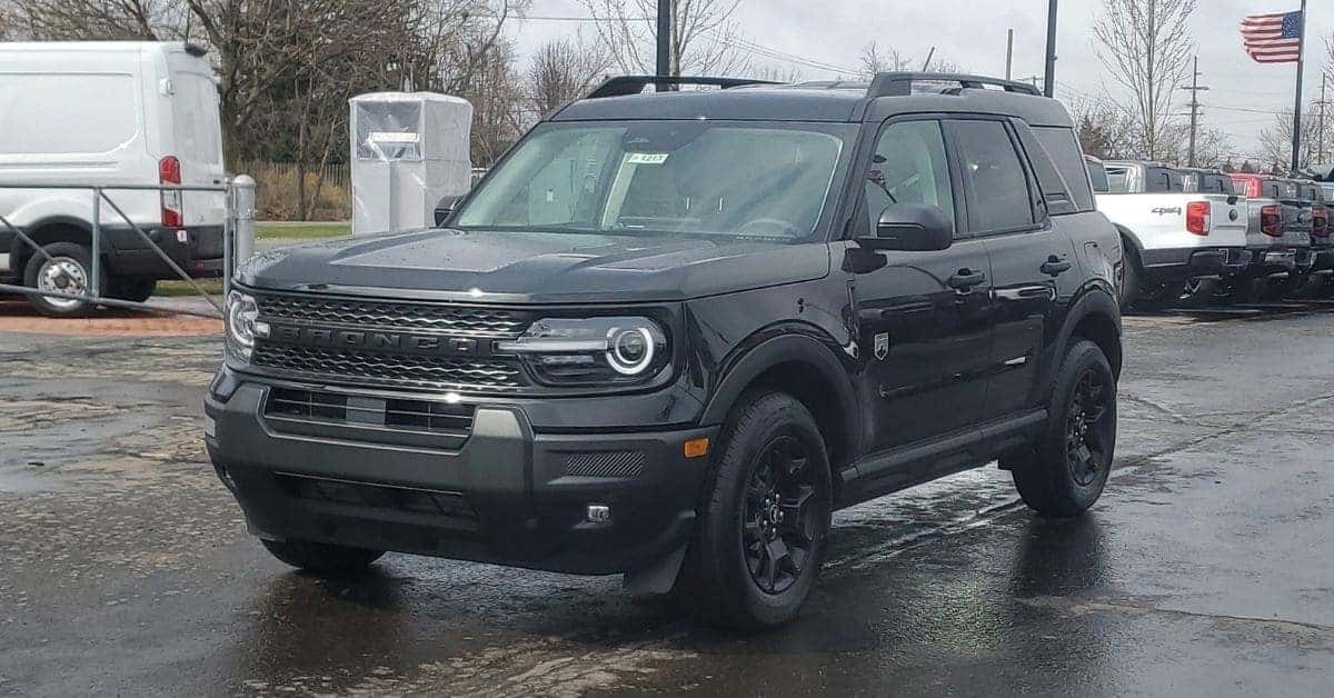 A black 2025 Ford Bronco Sport SUV parked on wet asphalt in a dealership lot, with a white cargo van and pickup trucks in the background, under a cloudy sky.