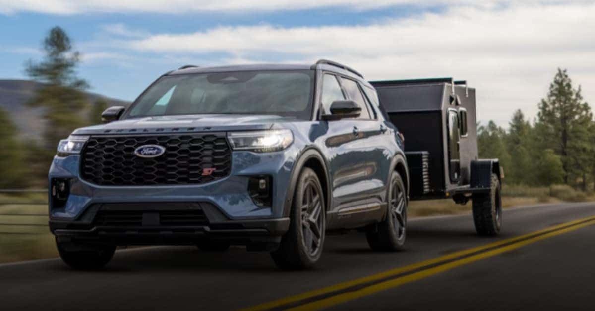 A blue 2026 Ford Explorer ST SUV towing a rugged black off-road trailer on a scenic paved road surrounded by rolling hills and pine trees under a partly cloudy sky, emphasizing its towing and adventure capabilities.