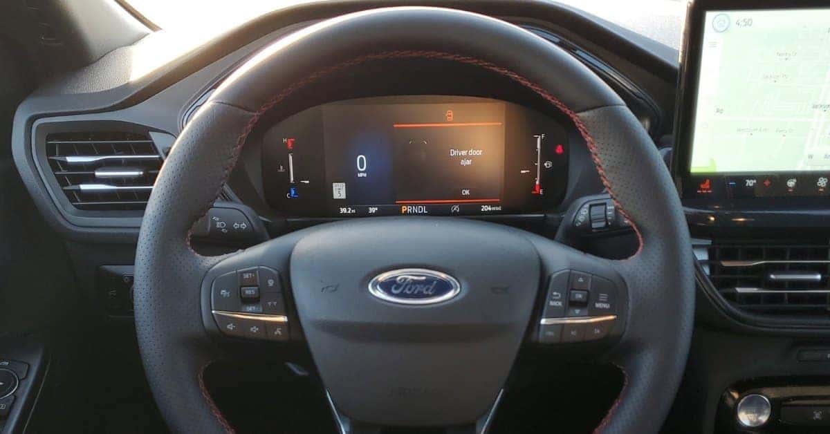 Interior view of a 2026 Ford Escape showcasing the digital dashboard with a speedometer reading 0 MPH, a 'Driver door ajar' warning, and an infotainment screen displaying a map and climate controls. The steering wheel features red stitching and multi-function buttons.