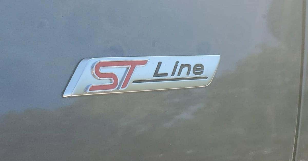 Close-up of a metallic 'ST Line' emblem on a gray 2026 Ford Escape surface. The emblem features bold red 'ST' letters and a sleek design, signifying a sporty trim level of the 2026 Ford Escape.