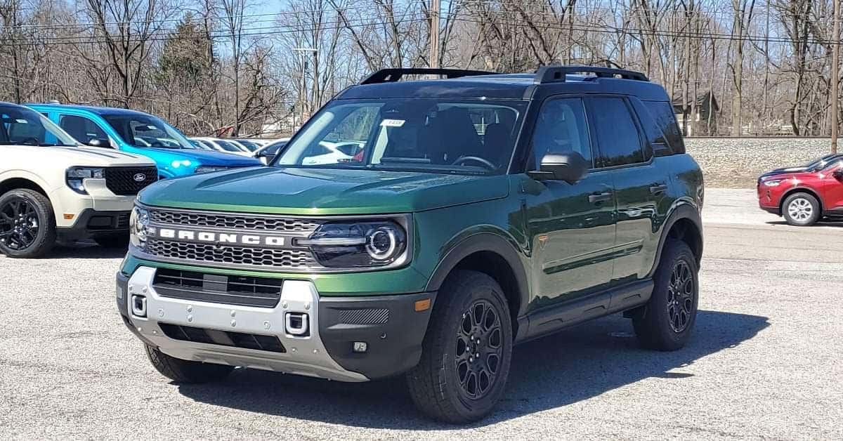 A green 2025 Ford Bronco Sport SUV parked at the Varsity Ford dealership lot on a sunny day, surrounded by other vehicles, including a beige and blue Bronco Sport, with bare trees in the background.