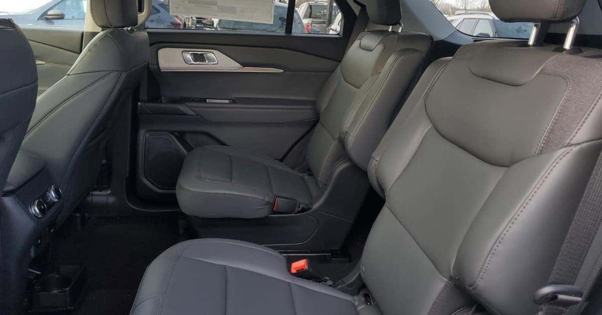 The interior of the 2026 Ford Explorer SUV showcases second-row captain's chairs with fabric accents and a partially visible third row, along with a rear console, door panel with metallic trim, and a view of a dealership lot through the windows.