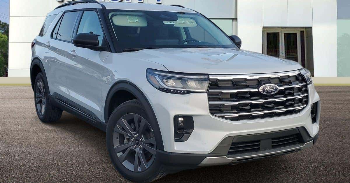 A light grey 2026 Ford Explorer SUV parked on an asphalt surface in front of Varsity Ford with large windows, showcasing its bold grille, sleek headlights, and rugged design elements like black fender flares and roof rails.