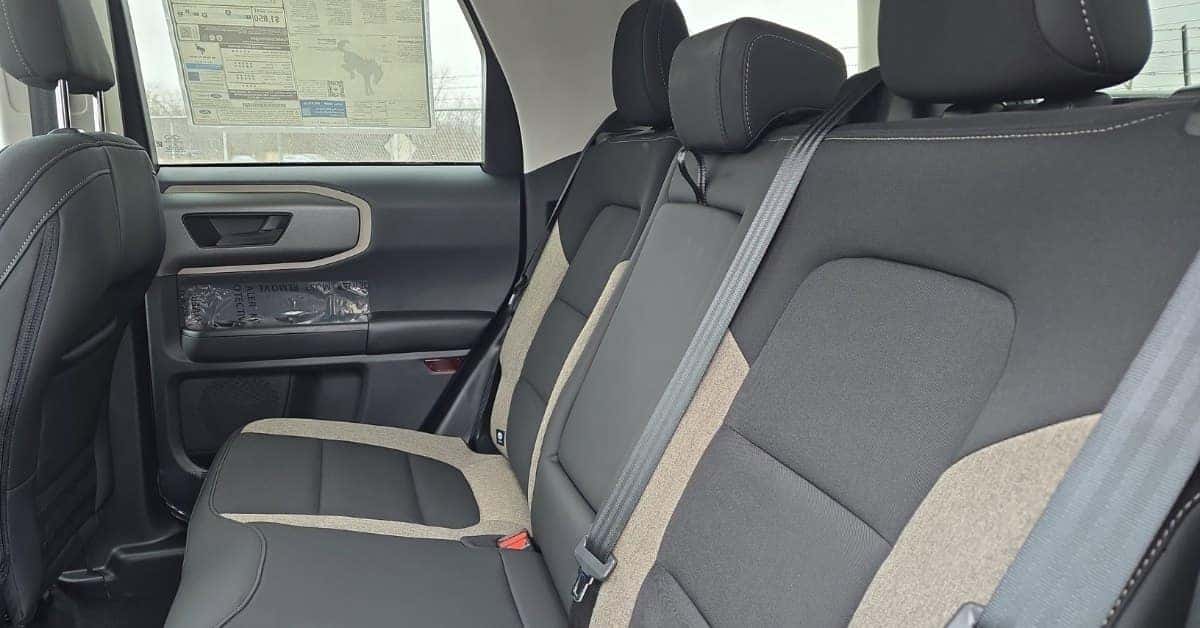 Interior of a new 2026 Ford Bronco Sport showcasing the rear passenger area with two-tone fabric seats, a protective plastic covering on the door panel, and a window sticker visible on the side window, indicating the car's new condition.
