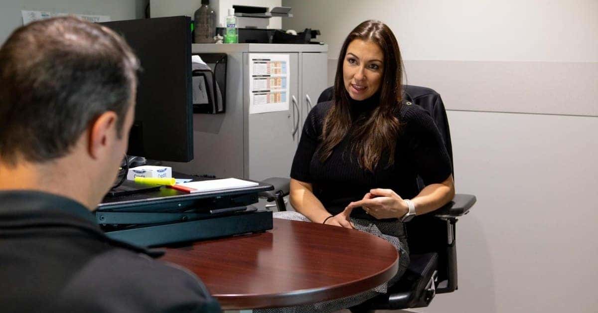 A professional discussion between a man and a woman in an office setting, seated at a round wooden table with a computer monitor, office supplies, and a filing cabinet in the background, suggesting a client-advisor interaction.