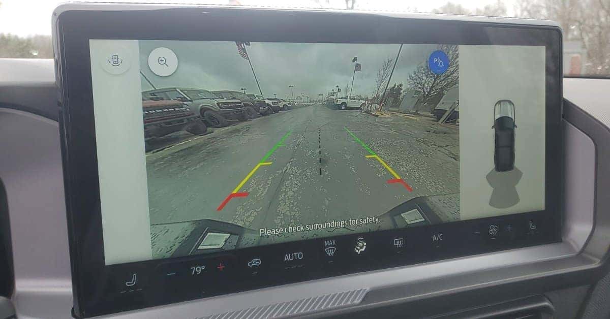 An in-car infotainment screen of the 2025 Ford Bronco Sport showing a rearview camera feed with dynamic guidance lines, parked vehicles in a dealership lot, and climate control settings displayed at the bottom.