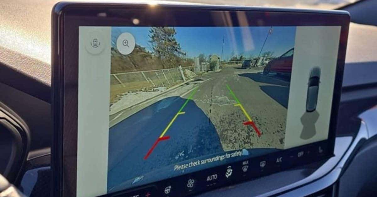 A 2026 Ford Escape infotainment screen displaying a rearview camera feed with dynamic parking guidelines. The feed shows a road with a sloped embankment on the left and a red car parked on the right. A top-down schematic view of the car is visible on the side panel, and the bottom bar includes climate control and safety icons.