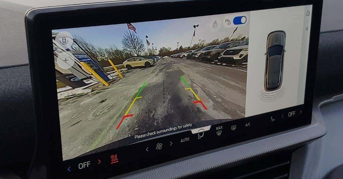 A 2026 Ford Explorer's infotainment screen showing a rearview camera feed with parking guidance lines, a split-screen overhead vehicle view, and various touch controls for climate and parking assist, set against a dealership parking lot with rows of vehicles.