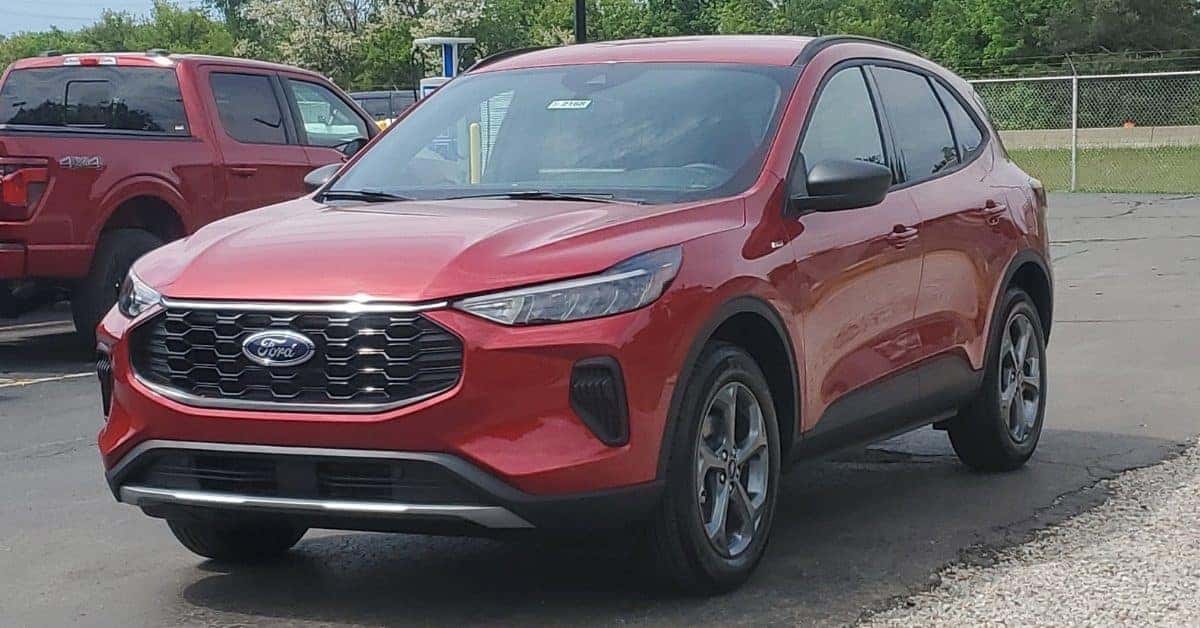 A red 2026 Ford Escape parked in the foreground with a red Ford F-150 pickup truck partially visible in the background. The vehicles are parked outdoors on a paved lot at Varsity Ford with trees and a chain-link fence in the background.