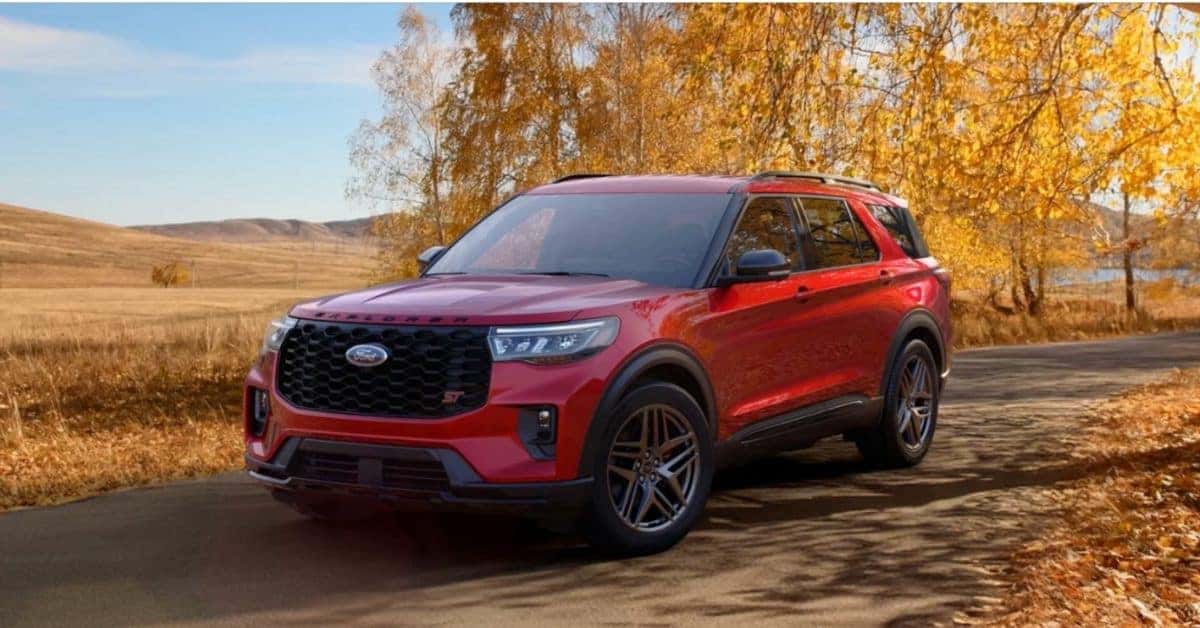A red 2026 Ford Explorer ST SUV parked on a curved asphalt road surrounded by vibrant autumn foliage and rolling golden hills. The vehicle's bold grille, "EXPLORER" lettering, and red "ST" badge are prominently displayed, with a clear blue sky and a reflective body of water in the background, emphasizing its adventurous appeal.