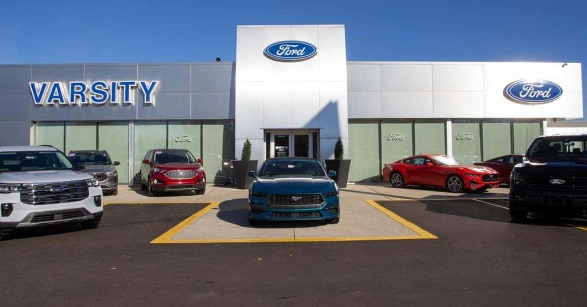 Exterior of the Varsity Ford dealership on a sunny day, featuring a modern facade with 'VARSITY' in blue letters, the Ford logo above the entrance, and several Ford vehicles, including SUVs, Mustangs, and a pickup truck, parked in the lot.