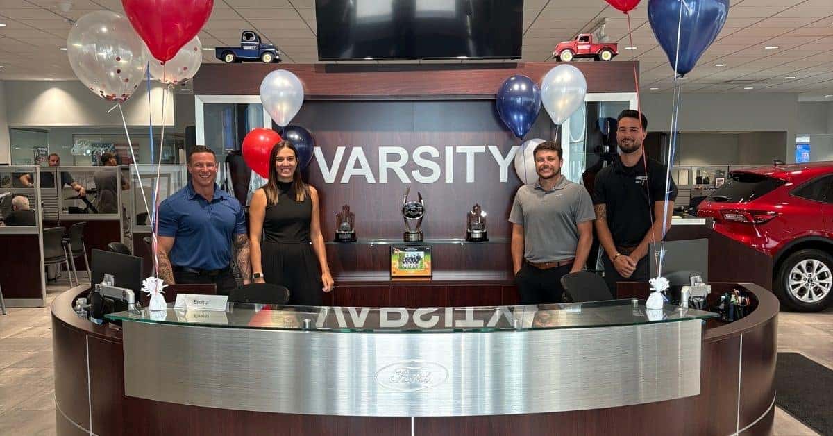 Four smiling employees stand behind a curved reception desk at Varsity Ford, featuring a metallic Ford logo. The background showcases "VARSITY" branding, trophies, and festive red, white, and blue balloons, creating a welcoming and celebratory atmosphere in the dealership.